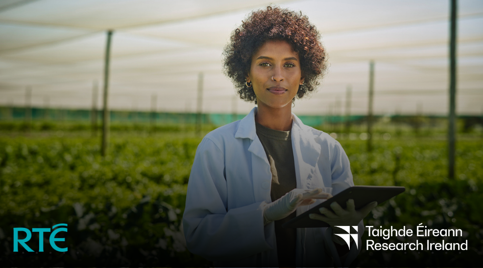 Woman wearing a white lab coat is holding a tablet device. There is fields of crops in the background. Research Ireland and RTÉ logos are on the image.